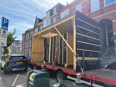 Een prefab element staat vastgesjord op een dieplader in de straat, ingepakt in zwarte folie en met houten schoren erin.