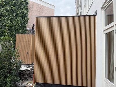 Een aanbouw met verticale houten latten in een achtertuin, met daarachter een kleiner bijgebouw in dezelfde latten.