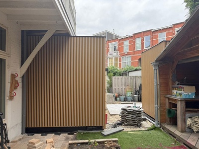 De aanbouw met houten latten vanuit de tuin gezien, met de woning links en een houten overkapping rechts.