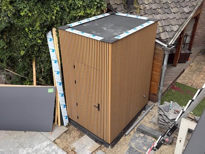 De afgeronde aanbouw van bovenaf: een plat dak met de randen nog afgeplakt, en een gevel van verticale houten latten.